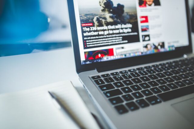Birou de redacție cu un laptop deschis, ziare împăturite și un carnet de notițe lângă o cană de cafea
