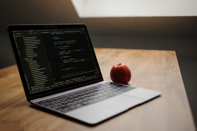 Telefon care afișează un cod de verificare, ținut în mână lângă un laptop deschis pe pagina de autentificare