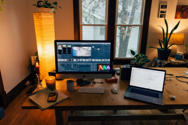 Birou cu un monitor așezat lângă o fereastră luminoasă, cu o lampă de birou și o plantă verde alături